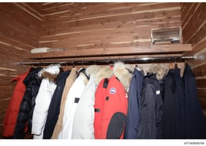 This photograph shows a collection of luxury winter coats and jackets hanging on a metal rod in what appears to be a closet or storage room with wooden ceiling and walls. The collection includes premium outerwear in red, black, white, and navy colors