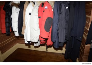 This photograph documents a collection of winter outerwear including puffy jackets, parkas, and coats in various colors (red, white, black, navy, tan) hanging on a rod. The items appear to be evidence collected during an investigation, photographed i