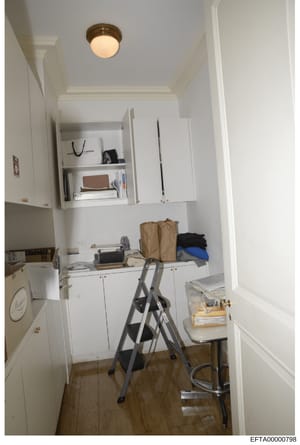 This is a photograph of a narrow, white-walled room with built-in cabinetry and storage shelving. The space appears to be used for storage and office work, containing various personal items, boxes, clothing, and supplies. The room features white pain