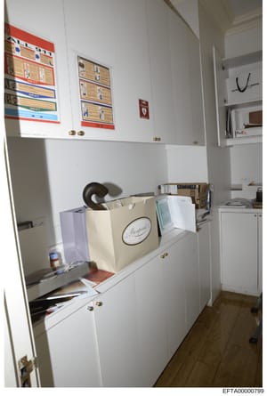 This photograph shows an interior office or workspace with white cabinetry, displaying educational materials and charts on the upper cabinet doors. The workspace contains various items including shopping bags, documents, and office supplies. The imag