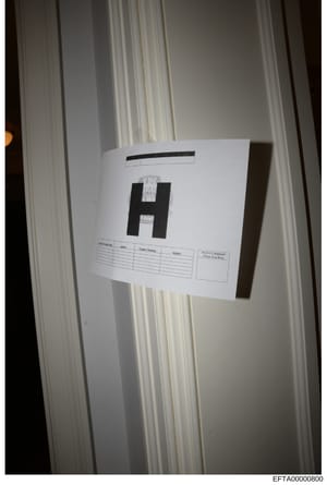 This is a photograph of evidence documentation showing a large black letter 'H' with an evidence label attached, positioned against white curtains or drapes. The label appears to be part of a formal evidence collection and cataloging procedure, with 