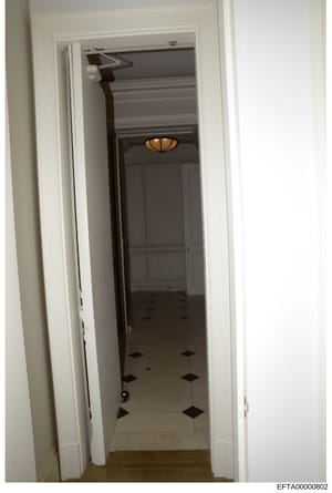 This is a photograph of an interior hallway, likely from one of Jeffrey Epstein's properties, showing a narrow corridor with white-painted walls and a distinctive black-and-white diamond-patterned floor. A dark wooden door is visible at the end of th