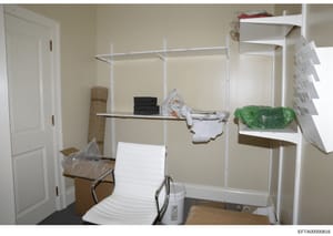 This photograph documents an interior room space containing white metal shelving units with various stored items including boxes, clothing, household goods, and other materials. The image appears to be evidence documentation from a property search or