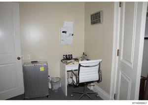 This is an evidence photograph showing an interior office or workspace, likely from one of Epstein's properties. The image captures a small office area with a desk, office chair, filing/mail station on the wall, a wheeled suitcase, waste basket, digi