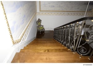 This is a photograph of an elegant interior staircase featuring ornate wrought-iron railings, gold-trimmed crown molding, and high-end decorative elements including an antique chair with green upholstery. The image appears to be documentation of prop