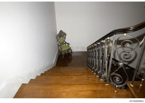 This photograph shows an interior staircase with hardwood flooring, white walls, and an ornate metal railing with decorative scrollwork. A period-style chair with a green seat and carved wooden frame is visible on the landing. The image appears to be