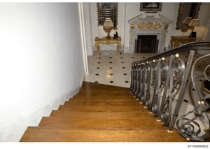 This photograph shows the interior of a luxury residence, specifically a staircase and foyer area featuring expensive period furnishings including decorative marble fireplaces, gilt mirrors, ornamental tables, and wall sconces. The image appears to b