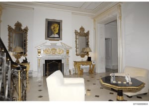 This photograph documents an elegantly furnished interior room featuring neoclassical design elements, including marble fireplace, ornate gold-leafed mirrors, decorative console tables, and fine art. The space appears to be part of a high-end residen