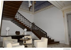 This is a photograph of an opulent interior space featuring a grand wooden staircase with ornate wrought iron railings, cream-colored upholstered seating, decorative sculptures, and detailed ceiling work with gold leaf trim. The architectural style a