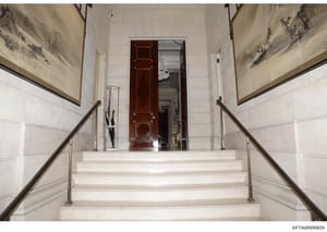 This photograph shows an elegant interior staircase with cream-colored marble or stone steps, metal handrails, and walls decorated with framed Asian landscape artwork. A dark red wooden door is visible at the top of the stairwell, with hallways exten