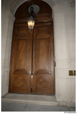 This photograph shows the exterior entrance to a property, featuring a pair of tall wooden doors with detailed paneling set within a rounded arch. The doors are constructed of dark wood with decorative hardware and are illuminated by a vintage-style 