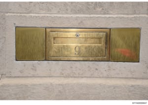 This is a photograph of a decorative brass mailbox mounted on a concrete or stone wall. The three-panel fixture features a central mail slot with the number '9' displayed, flanked by two blank brass panels. The mailbox shows signs of age and patina, 
