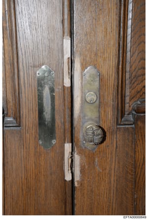 This is a close-up photograph of a pair of wooden double doors featuring decorative brass or bronze hardware. The doors appear to be interior doors from a high-end residence or property, with dark wood paneling and ornate door plates on each panel. T
