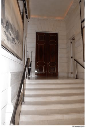 This is a photograph of an elegant interior staircase and hallway featuring light-colored marble or stone flooring, white walls with paneling, a dark wood double door with brass hardware at the landing, and modern metal handrails. The space appears t