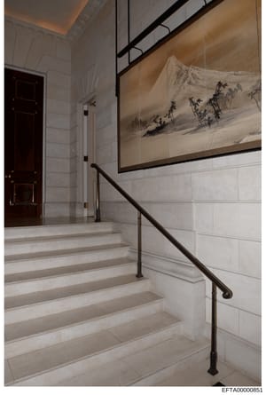 This photograph shows the interior of a stairwell in what appears to be a luxury residential property. The image captures a contemporary design space featuring white marble or stone steps, a dark metal handrail, white subway tile walls, and a large f