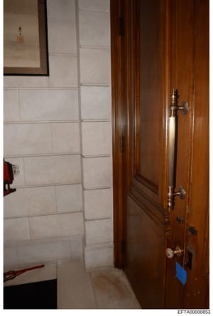 This is an evidence photograph showing the interior of a residential space, featuring a wooden door with brass handles on the right side and cream/beige tile wall paneling on the left. A framed item hangs on the upper left wall. The photo appears to 