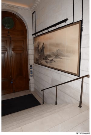 This is a photograph of an interior hallway or entryway featuring classical architectural elements and decorative artwork. The space shows a wooden arched door with a circular window, white tile walls, and a large framed landscape painting depicting 