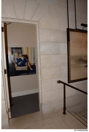 This is an evidence photograph showing an interior hallway or corridor in what appears to be an Epstein property. The image depicts a cream-colored tiled wall, a white-framed doorway, and a framed portrait of a woman in a blue dress displayed on the 