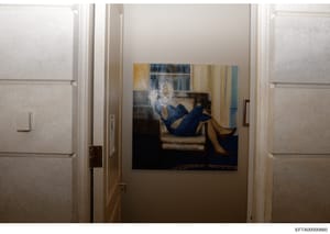 This is a photograph of an interior hallway or corridor showing a framed artwork hung on a cream-colored wall. The artwork depicts a figure in dark clothing reclining on furniture. The space features white trim, doors on either side, and warm lightin
