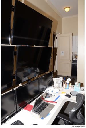 This photograph shows an interior office space with multiple computer monitors mounted on a wall, a work desk with a laptop and various office supplies, and doorways leading to other rooms. The image appears to be evidence documentation from a proper