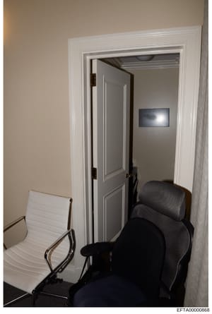 This is a photograph of an interior office or waiting area showing a small room with modern furnishings. The image depicts two chairs (one white, one black mesh office chair) positioned in front of an open doorway leading to an adjacent room with gra