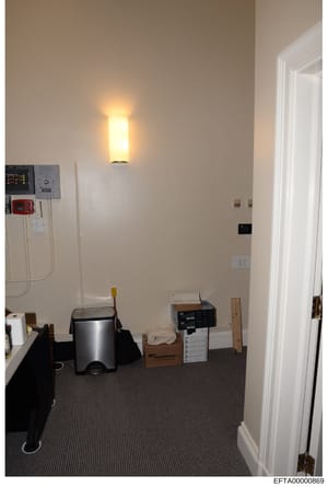 This is a photograph of an interior room showing various building systems and equipment installations. The image depicts security/control panels on the left wall, a fire alarm box, lighting fixtures, electrical outlets, and miscellaneous equipment an