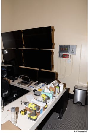 This photograph shows a control room or monitoring station setup with six large black display screens arranged in a grid pattern, mounted on a wooden frame. Below are laptops, various electronic monitoring equipment, and tools scattered on a desk. Th