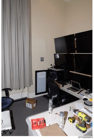 This is a photograph of an interior office or workspace showing a computer workstation with multiple monitors, desk equipment, tools, and furnishings. The image appears to be evidence documentation from the investigation, capturing the physical setup