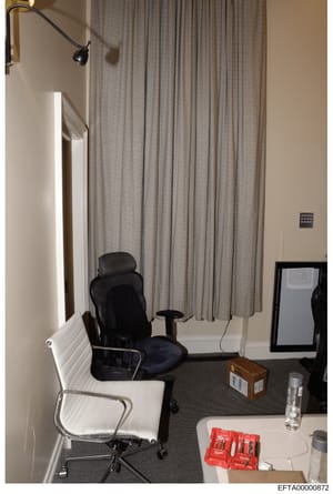 This is a photograph of an interior office or workspace showing a desk area with computer equipment, office chairs, and furnishings. The image appears to be evidence documentation from a property search or seizure, showing the layout and contents of 