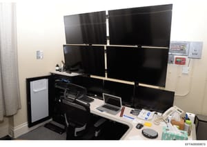 This photograph shows a sophisticated multi-monitor workstation setup with six display screens arranged in a 3x2 configuration, a laptop, and various office equipment. The setup appears designed for monitoring or surveillance purposes, with control p