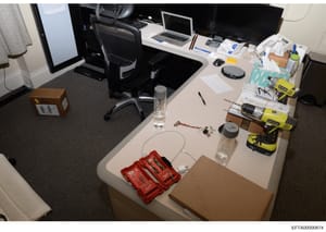 This is a photograph of an office workspace showing an organized desk with computer equipment, power tools, storage boxes, and miscellaneous items. The image appears to be evidence documentation from the investigation, possibly showing a workspace us