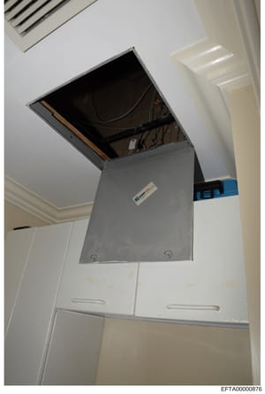 This is a photograph of a ceiling-mounted access panel or electrical box in what appears to be a residential interior space. The image shows an open gray metal panel with visible wiring and equipment inside, mounted flush with a white ceiling. The ph
