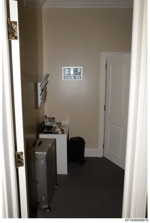 This is a photograph of a small interior room or closet space, showing a digital clock on the wall, luggage, and various personal items on a white shelf. The image appears to be evidence documentation from the investigation, with a Bates stamp indica