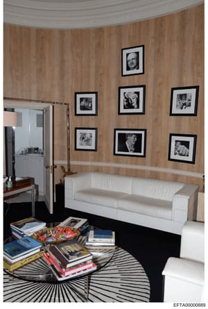 This is an evidence photograph documenting an interior room, likely from one of Jeffrey Epstein's properties. The room features a white sofa, wood-paneled walls, and multiple framed black-and-white portrait photographs displayed on the wall. A coffee
