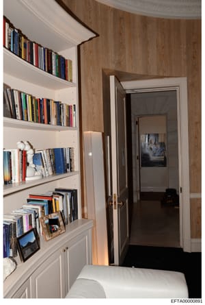 This is a photograph of an interior room featuring extensive built-in bookshelves with multiple shelves containing numerous books, decorative items, and framed photographs. The room has wood-paneled walls, white trim, and a doorway leading to an adja