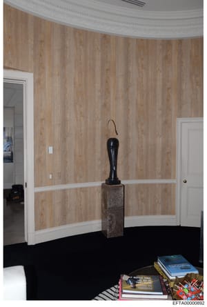 This is a photograph of an interior room featuring curved wood-grain wallpaper walls with white trim, a black sculpture on a stone pedestal, and furnishings including books and decorative items. The image appears to be from a property owned or contro