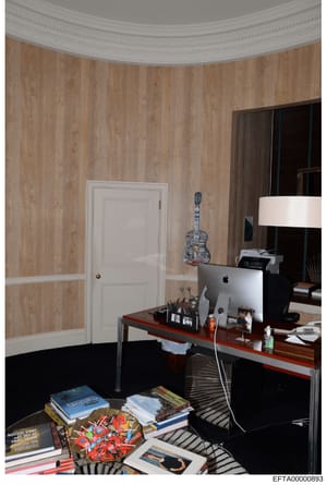 This is a photograph of an interior room, likely a bedroom or office, showing a desk workspace with a computer monitor, various personal items, books, and decorative elements. The room features wood-grain wallpaper, a curved ceiling detail, and a whi