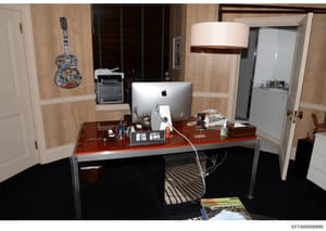 This photograph shows an interior office or study space featuring a wooden desk with computer equipment, decorative artwork, and personal items. The room contains wood-paneled walls, white doors, a framed guitar artwork, and various furnishings. The 