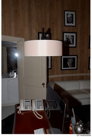 This photograph documents an interior room space, likely from one of Jeffrey Epstein's properties, showing multiple telephone systems, framed portrait artwork on wood-paneled walls, and furniture including a lamp and sofa. The presence of multiple ph