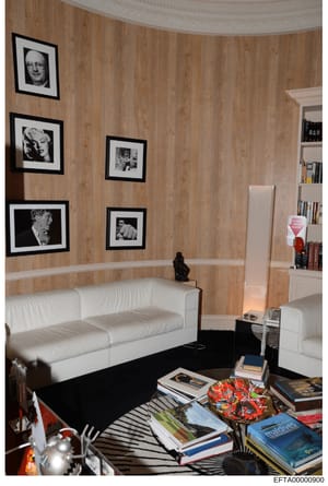 This photograph documents an interior room, likely from one of Jeffrey Epstein's properties, featuring wood-paneled walls with framed black-and-white portrait photographs, white upholstered seating, built-in shelving, and various decorative items and