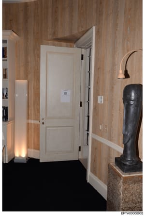 This is an evidence photograph from the Epstein investigation showing an interior room featuring wood-paneled walls, a white door marked with letter 'B', decorative sculptures including a black abstract bust, and interior lighting. The photograph doc