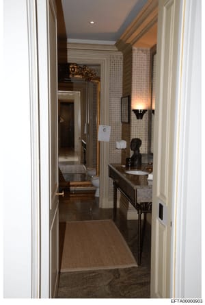 This is a photograph of an upscale, contemporary bathroom taken as evidence documentation. The image shows a hallway view into a luxuriously appointed bathroom with high-end finishes, including dual vanities, decorative wall sconces, patterned wallpa