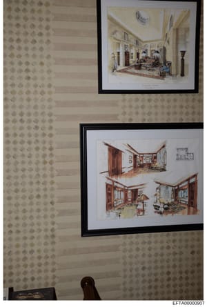 This photograph shows two framed architectural interior design renderings displayed on a patterned wallpapered wall. The renderings appear to be professional design sketches depicting elegant residential rooms with detailed furniture placement, color