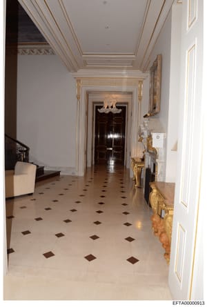This photograph depicts an elegantly appointed hallway featuring classical architectural elements including ornate gold leaf trim, decorative ceiling with geometric patterns, marble flooring with inlaid dark stone diamonds, and ornamental sculptures.