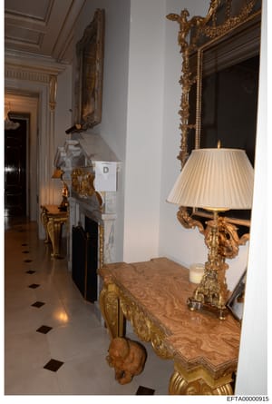 This is a photograph of an ornate interior hallway, likely from one of Jeffrey Epstein's residences, showing luxury furnishings and décor. The image depicts a marble-floored corridor with elaborate gold-leaf mirrors, a wooden console table with ornam