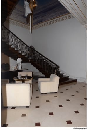 This is an evidence photograph showing the interior of a luxury residence, specifically a foyer or entry hall featuring a grand wooden staircase with ornate iron railings, decorative ceiling with gold leaf trim, cream-colored seating furniture, and a