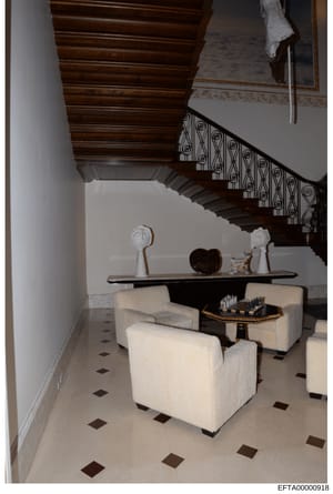 This is an evidence photograph documenting the interior of a high-end residential property, showing an elegant foyer or sitting area with a grand wooden staircase, decorative console table, white upholstered furniture, and ornate architectural detail