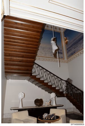This photograph documents the interior of a high-end residence, showing an ornate wooden staircase with decorative iron railings, coffered ceiling details, and a curated art collection including white sculptural busts and a bronze heart sculpture on 