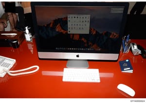 This is an evidence photograph showing a desk setup with an Apple iMac computer, wireless keyboard and mouse, office supplies, and a U.S. passport. The photograph documents the layout and contents of a workspace, likely seized or photographed during 