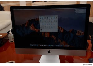 This is a photograph of an Apple iMac computer displaying a macOS desktop with a file manager window open showing various folders and files. The computer is positioned on an orange/red desk surface in what appears to be an interior office or resident